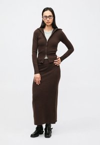 Rjav pleten jopič z zadrgo spredaj, kombiniran z ujemajočo se dolgo krilo. Oblečen s svetlo majico in črnimi gležnjarji. Gladek tekstura.