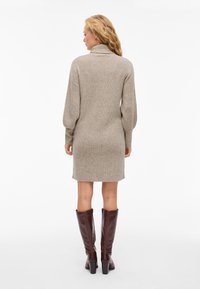 Smėlio spalvos megztinė su iškiliu apykakle ir ilgomis rankovėmis, baigianti virš kelių, derinama su ruda odine aukštakulnėmis batais iki kelių.