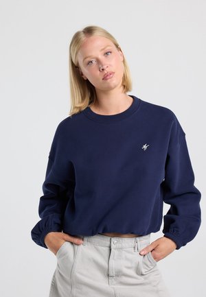 Blondine wearing a navy blue cropped sweatshirt with elastic cuffs and light beige trousers, standing with hands in pockets.