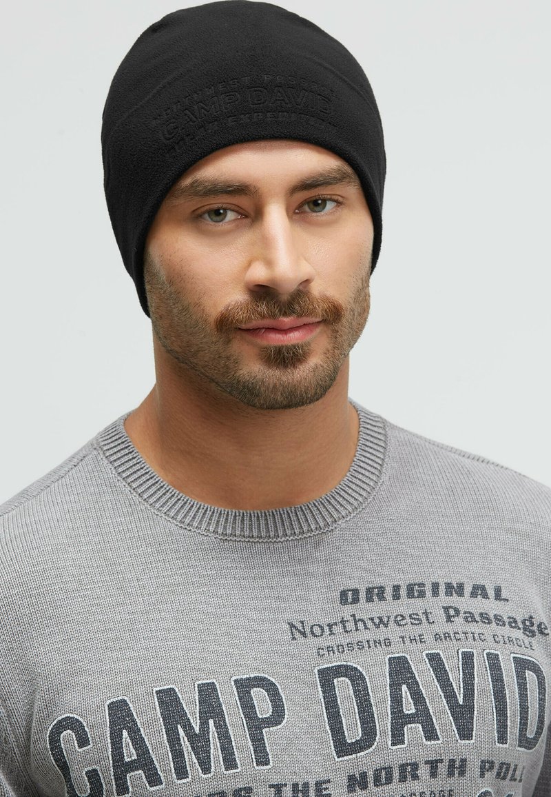 Black knit beanie with a textured finish and embossed logo. Worn with a light gray sweater featuring printed text and a ribbed neckline.
