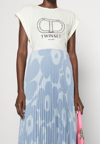 Vrouw in een witte Twinset Milano t-shirt en een blauwe bloemenplissé rok, met een felroze handtas naast zich.