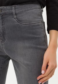 Los jeans de mezclilla grises presentan un diseño ajustado, un estilo clásico de cinco bolsillos y sutiles acentos de costura. La textura es suave con un ligero brillo.