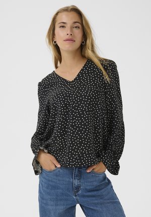 Blusa nera a pois con scollatura a V, maniche lunghe e polsini arricciati, realizzata in un tessuto leggero. Abbinata a jeans blu.