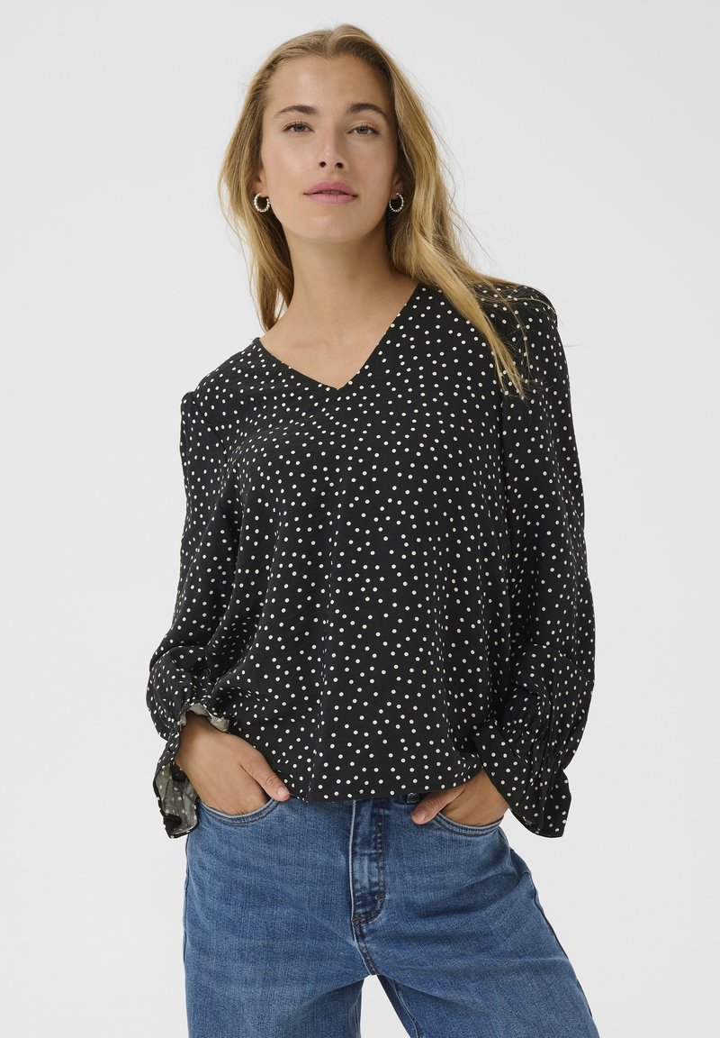 Blusa nera a pois con scollatura a V, maniche lunghe e polsini arricciati, realizzata in un tessuto leggero. Abbinata a jeans blu.