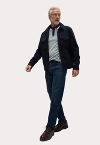 Marineblå jacka med två bröstfickor över en ljusblå polo, kombinerad med mörk denimjeans och bruna skor. Man går med självförtroende.