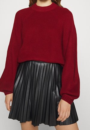 Femme portant un pull rouge en maille avec des manches bouffantes, associé à une mini-jupe plissée en cuir noir.