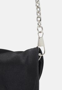 Czarna torba z tkaniny o miękkiej fakturze, z srebrnym łańcuszkiem i metalowym zapięciem, podkreślająca nowoczesny, minimalistyczny design.