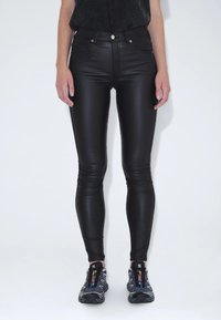 Černé legíny z umělé kůže s hladkým povrchem, vysokým pasem a úzkým střihem, doplněné šedými sportovními botami s barevnými akcenty.