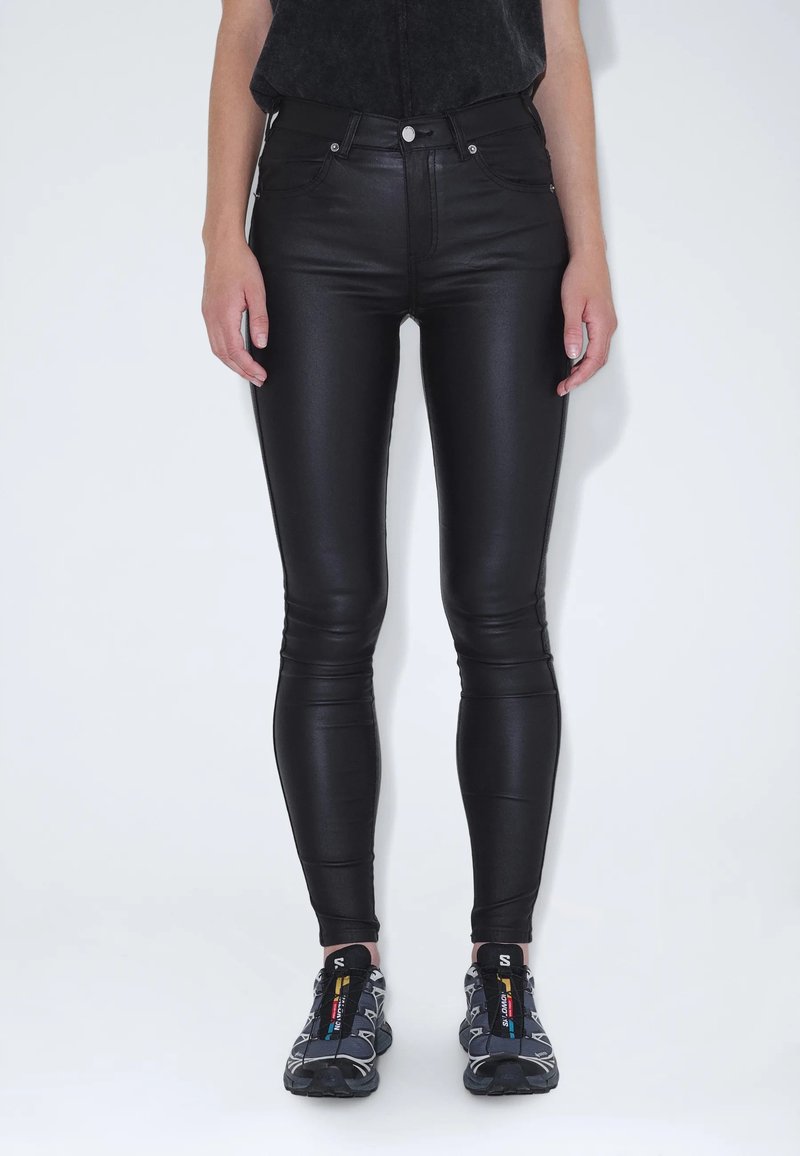 Černé legíny z umělé kůže s hladkým povrchem, vysokým pasem a úzkým střihem, doplněné šedými sportovními botami s barevnými akcenty.