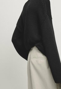 Musta neulepusero, jossa on löysä istuvuus ja näkyvä rakenne, yhdistettynä vaaleanbeigeihin housuihin, joissa on takatasku.