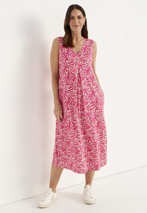 Frau steht vor einer weißen Wand, trägt ein rosa ärmelloses Midikleid mit Blumenmuster und weiße Turnschuhe, lächelt mit den Händen in den Taschen.