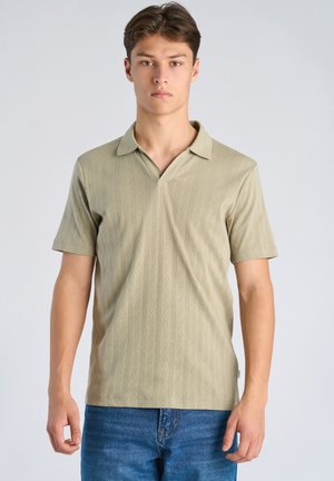 Giovane uomo fermo in piedi, indossa una polo beige a maniche corte senza bottoni e jeans blu, di fronte a uno sfondo semplice.