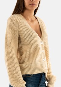 Cárdigan de punto en color beige claro con escote en V, que presenta grandes botones blancos y una textura acanalada. Ajuste suave y relajado con mangas largas.