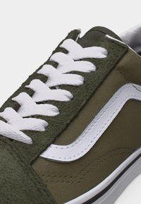 Baskets basses en daim vert olive et toile, avec une bande latérale blanche ondulée et des lacets blancs épais sur une semelle blanche.