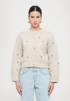 Beige strikket cardigan med rund halsudskæring, diamantmønster, broderede jordbær og bier samt frontknapper. Bæres over lyseblå jeans.