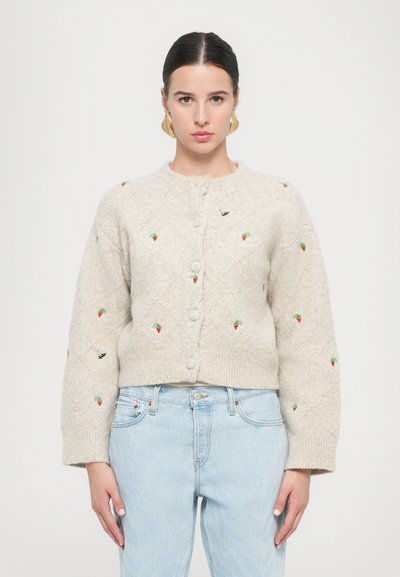 Beige gestrickte Strickjacke mit runder Ausschnitt, Rautenmuster, bestickten Erdbeeren und Bienen sowie frontalen Knöpfen. Getragen über hellblauen Jeans.
