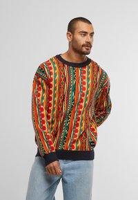 Kleurrijke gebreide trui met rode, gele, groene en blauwe patronen, voorzien van een geribbelde marineblauwe halslijn en mouwen, gecombineerd met lichtblauwe jeans.