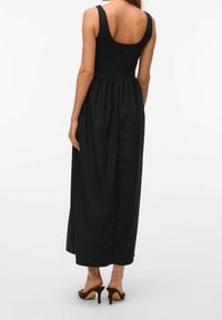 Un vestido maxi negro sin mangas con un corpete ajustado y una falda acampanada, de tela lisa y con un diseño de espalda en forma de barco. Llevado con unos zapatos de tacón alto marrones.