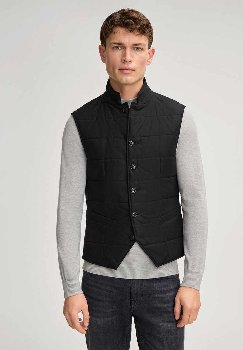 Czarna pikowana kamizelka z wysokim kołnierzem, zapinana na guziki z przodu, z bocznymi kieszeniami, noszona na jasnoszarym swetrze i zestawiona z ciemnymi dżinsami.