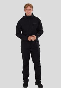 Ung man iklädd svart zip-up hoodies, svarta byxor med sidofickor och svarta snörskor, stående mot en enhetlig bakgrund.