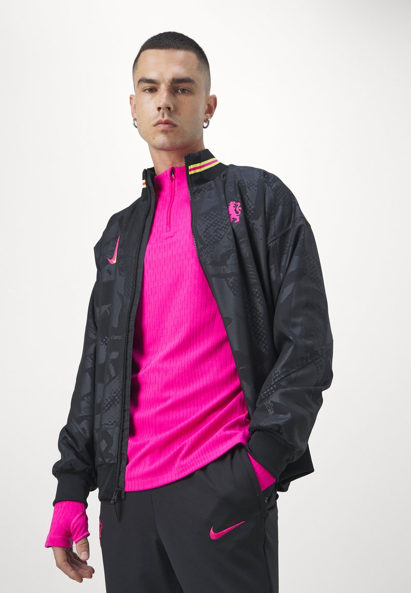 Giacca nera con zip e texture a motivo, con accenti neon, abbinata a un top fucsia con collo alto e pantaloni neri con logo.