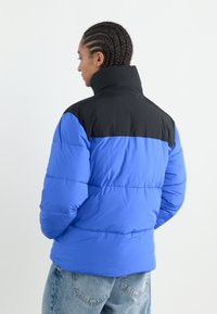 Puffervest med blå krop og sort krave. Fremstillet af letvægtsstof med quiltet tekstur og høj hals for varme.
