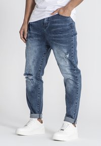 Pantalones vaqueros de denim azul con un ajuste relajado, que presentan áreas desgastadas en la rodilla y dobladillo acampanado, combinados con zapatillas blancas.