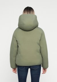 Giacca imbottita verde con un ampio cappuccio, caratterizzata da una texture liscia e polsini a costine, indossata su jeans di denim scuro. Vista da dietro.