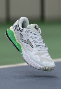Baskets blanches de sport avec un cuir texturé, accent de semelle extérieure verte et éléments de design noirs. Présente un laçage et des panneaux en mesh.