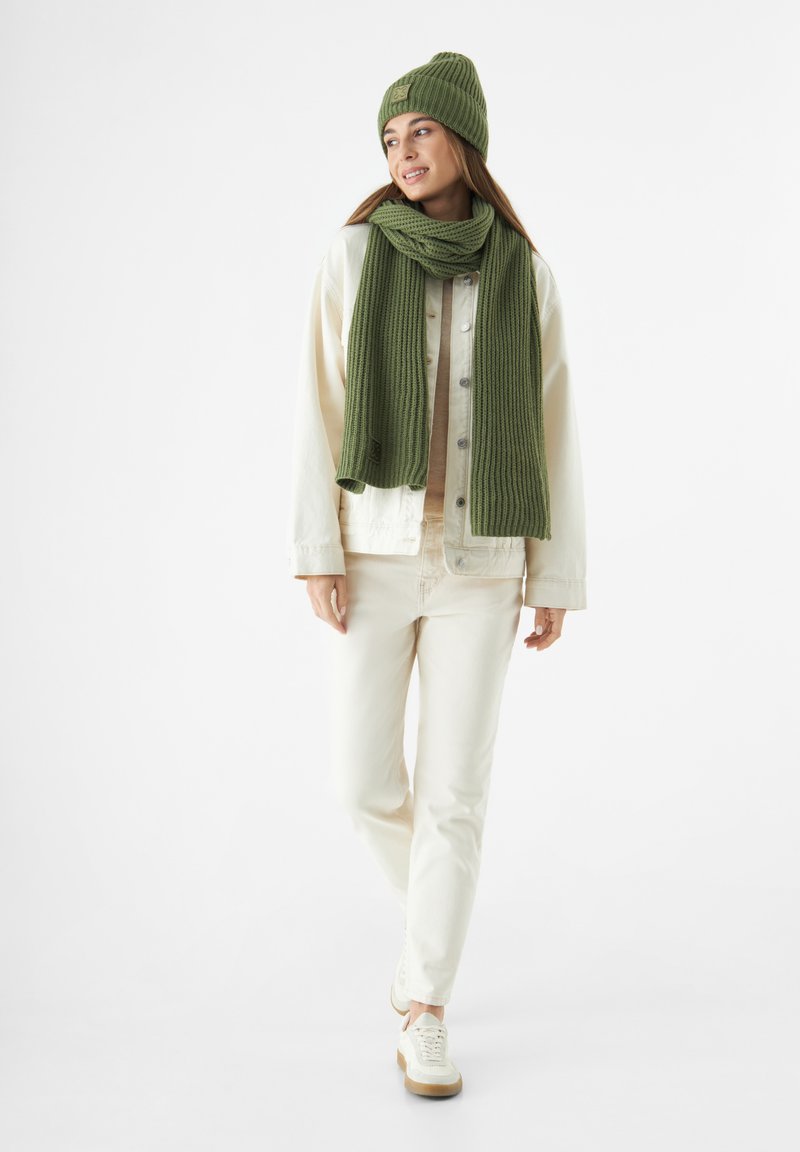 Groene gebreide beanie en sjaal set met ribbels. Het model draagt een crèmekleurige spijkerjack en strakke witte broek met witte sneakers.