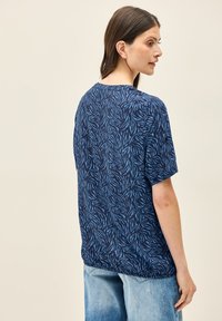 T-shirt blu a maniche corte con un motivo di foglie su uno sfondo blu navy scuro, realizzato in un tessuto morbido con una vestibilità rilassata, abbinato a jeans chiari.