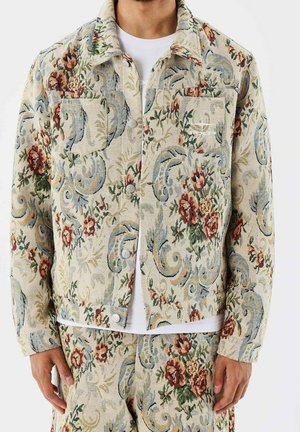 Homme portant une veste et un pantalon beige assortis avec des motifs floraux et cachemire dans des tons rouges, verts, bleus et crème.