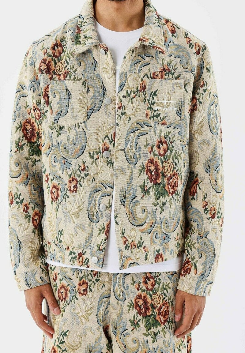 Homme portant une veste et un pantalon beige assortis avec des motifs floraux et cachemire dans des tons rouges, verts, bleus et crème.