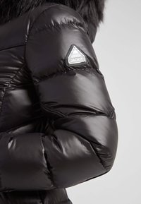 Chaqueta de plumas negra con acabado brillante, diseño acolchado y parche triangular con el texto "JACKIT" en la manga izquierda.