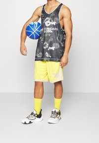 Black mesh basketball tank top with white graphic, paired with yellow shorts featuring a light beige accent. Holds a blue basketball, wears white sneakers.