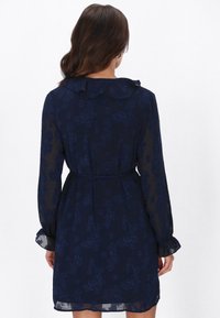 Tengerészkék virágos ruha csipkeszerű mintázattal, hosszú ujjakkal, fodros gallérral és testre simuló derékkal, térd fölött végződve.