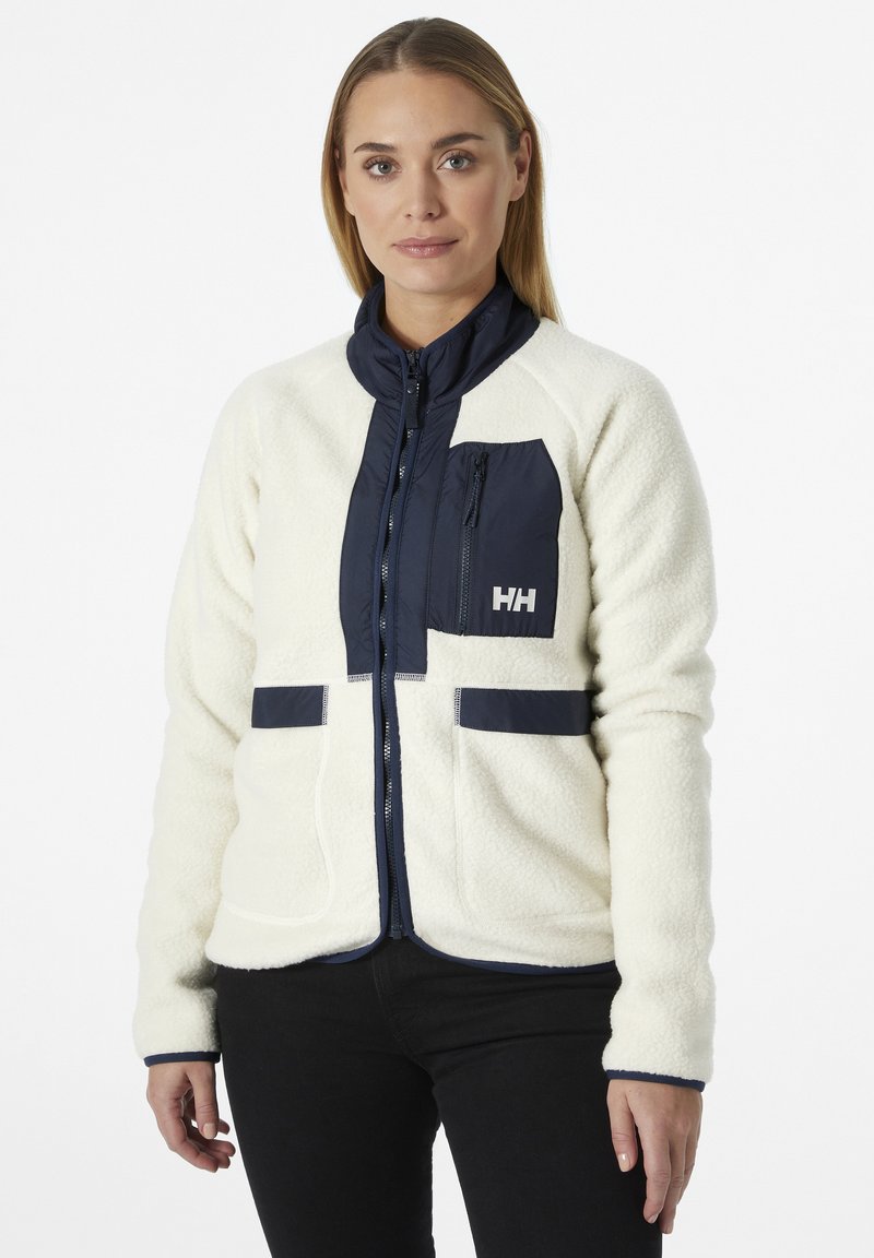 Chaqueta de forro polar en color crema con detalles en azul marino. Cuenta con cierre de cremallera, cuello alto y dos bolsillos frontales. Superficie texturizada con paneles en contraste.