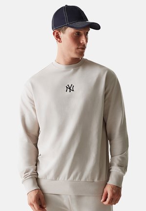 Hellgrauer Sweatshirt mit rundem Ausschnitt und gerippten Bündchen, versehen mit einem kleinen schwarzen gestickten Logo auf der Brust. Getragen mit einer marineblauen Mütze.