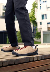 Beige outdoor shoes with red laces, rubber black soles, and gray accents. Worn with black pants while standing on wooden slats.