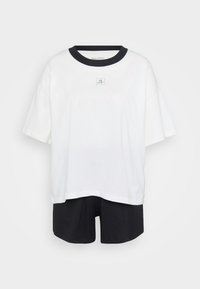 Weißes oversized T-Shirt mit schwarzem Kragen und kleinem Frontlogo, kombiniert mit schwarzen Shorts vor einem schlichten, hellen Hintergrund.