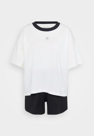 Valkoinen ylipitkä T-paita, jossa on musta kaulus ja pieni etulogo, yhdistettynä mustiin shortseihin tasaisella vaalealla taustalla.