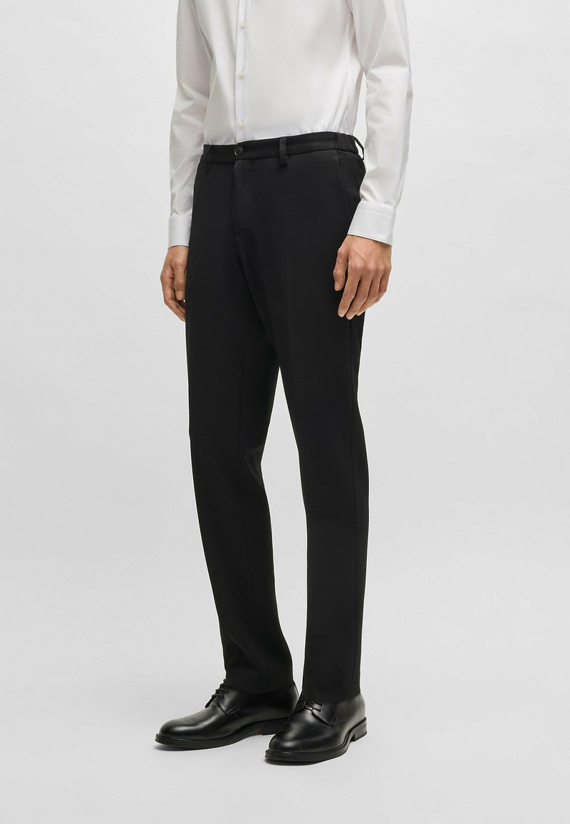 Pantaloni neri su misura realizzati in tessuto liscio, design a gamba dritta, con passanti per cintura e chiusura con bottone, abbinati a una camicia bianca e scarpe nere.