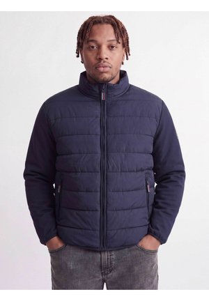 Homme aux cheveux tressés portant une veste matelassée bleu marine et un jean gris délavé, debout les mains dans les poches devant un fond uni.