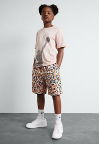 Camiseta de manga corta color rosa claro con un diseño gráfico, combinada con pantalones cortos de patrones coloridos. El calzado consiste en zapatillas blancas y calcetines.