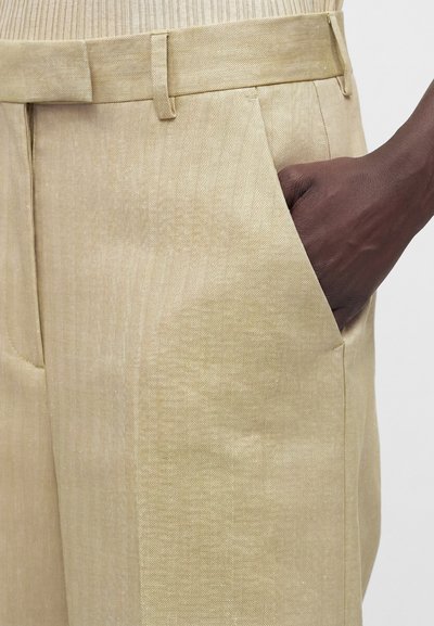 Χέρι με σκούρο δέρμα που ακουμπά στην τσέπη μιας μπεζ παντελόνας με σχέδιο ψαροκόκαλο και θηλιές για ζώνη.