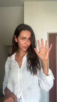 White linen shirt with a relaxed fit, featuring a small embroidered logo. Long, dark hair; hand raised in a greeting gesture. Light background.