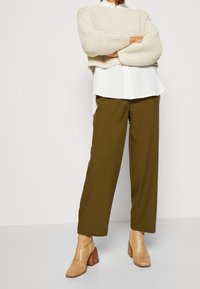 Pull en maille beige clair, chemise blanche en dessous, pantalon large vert olive, et bottines texturées beige avec un talon carré.