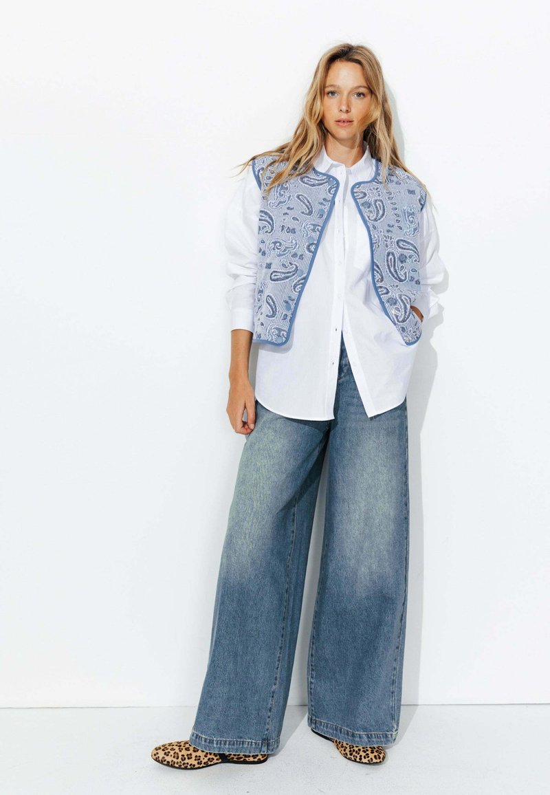 Gilet en denim bleu clair avec des motifs paisley, porté sur une chemise blanche et associé à un jean bleu délavé à jambes larges et des ballerines imprimées léopard.