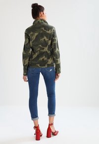 Bunda s maskáčovým vzorem vyrobená z bavlněné směsi, s límcem, staženým pasem a kombinovaná s úzkými modrými džíny a červenými robustními podpatky.