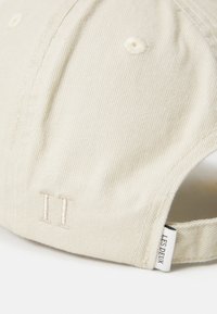 Nærbilde av en beige baseballcaps med brodert "II" og et hvitt stoffmerke som leser "LES DEUX" nær den justerbare stroppen.
